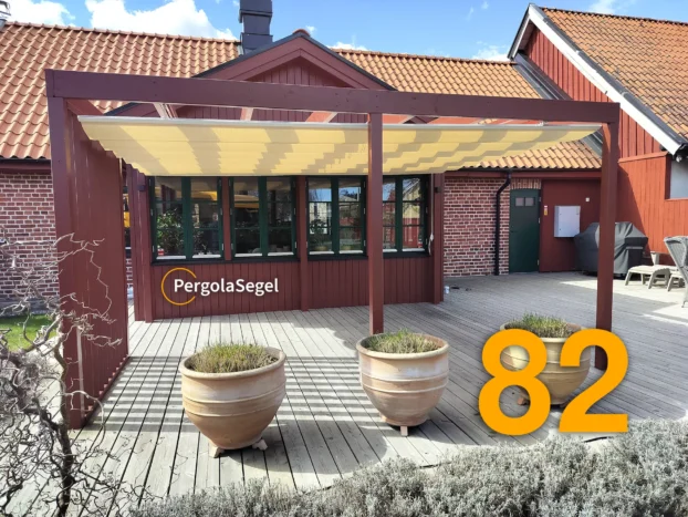 Faluröd pergola med beige segeltak framför husets fönster, sedd från trädgården i Åhus