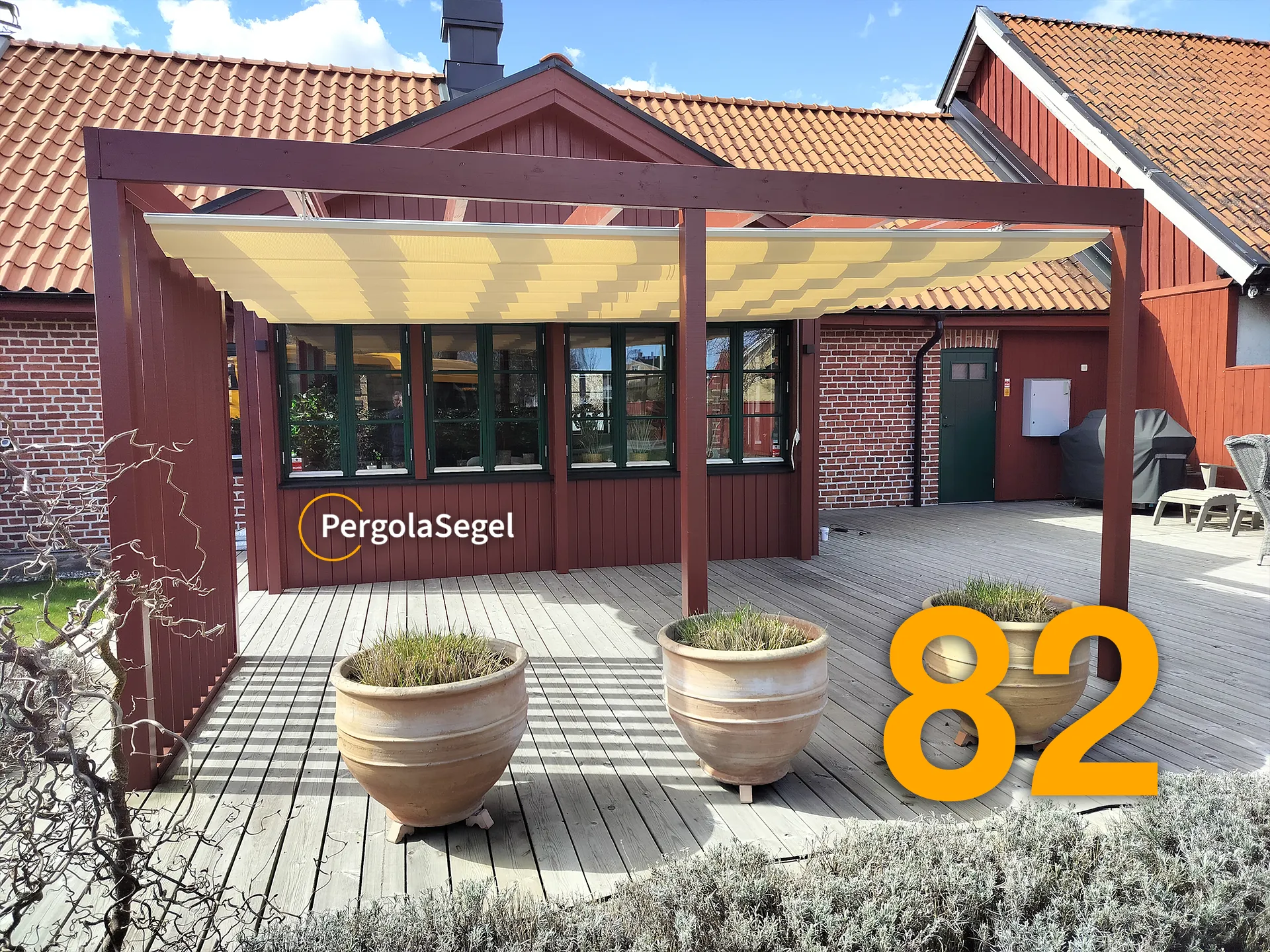 Faluröd pergola med beige segeltak framför husets fönster, sedd från trädgården i Åhus