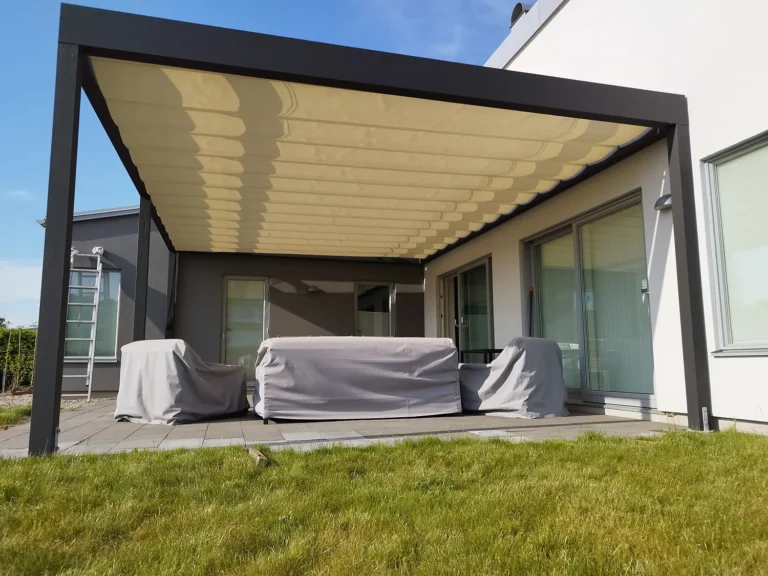 Svart väggmonterad pergola med beige pergolamarkis som öppnas från sidan över en skyddad uteplats